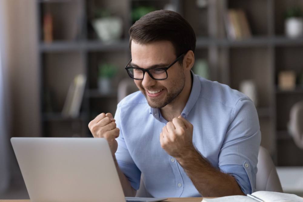 freelancerwinningabid Just won a bid on a freelancer site. young man with glasses sitting in front of a laptop computer looking happy.